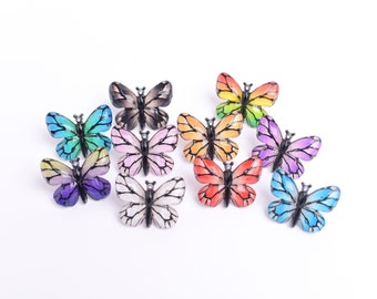 May include: A collection of colorful butterfly-shaped decorative pins. Each pin features a unique color scheme, including shades of blue, green, purple, pink, orange, and black. The pins are arranged on a white surface, showcasing their vibrant designs.