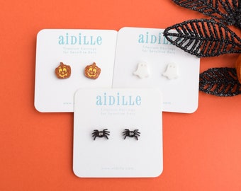 May include: Three pairs of Halloween-themed earrings on white cards against an orange background. The earrings feature pumpkin, ghost, and spider designs. The cards display the brand name "aidille" and the text "Titanium Earrings for Sensitive Ears."
