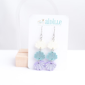 May include: A pair of silver dangle earrings with three layered seashells. The top seashells are white, the middle seashells are light blue with glitter, and the bottom seashells are purple with glitter. The earrings are on a white card with the text "aidille handmade accessories for a stylish home".