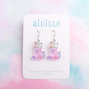May include: A pair of iridescent cat-shaped earrings with titanium ear wires. The earrings are on a white card with the text "aidille" and "Titanium Ear Wires for Sensitive Ears" and "www.aidille.com".