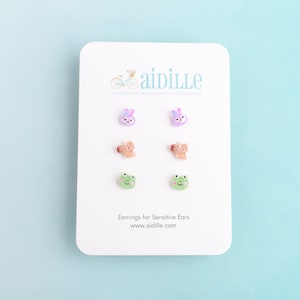 May include: A set of six small, colorful earrings featuring a bunny, a bear, and a frog. The earrings are displayed on a white card with the text "aidille" and "Earrings for Sensitive Ears".