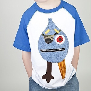 May include: A white and blue raglan sleeve t-shirt with a quirky blue monster design, including a red eye, camouflage patch, and a brown beak. The shirt is worn by a young person.