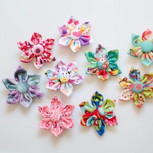 May include: A collection of ten fabric flowers, each featuring six petals and a central fabric-covered button. The flowers display various colorful patterns, including floral, geometric, and animal prints in shades of pink, blue, green, and yellow.