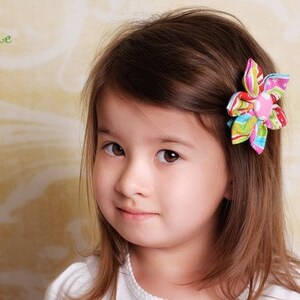 May include: A fabric hair clip featuring a multi-colored floral design with stripes, dots, and swirls in shades of pink, green, blue, and yellow, centered with a solid pink button. The word Aidille is printed in green in the upper left corner.