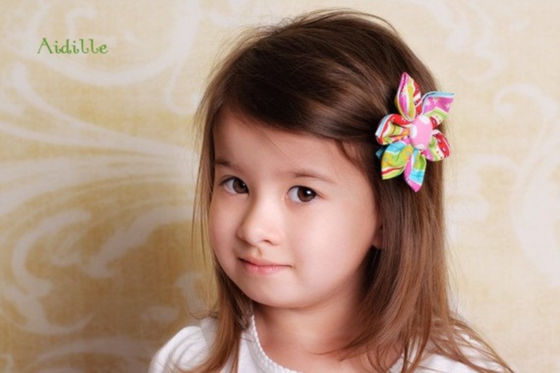 May include: A fabric hair clip featuring a multi-colored floral design with stripes, dots, and swirls in shades of pink, green, blue, and yellow, centered with a solid pink button. The word Aidille is printed in green in the upper left corner.