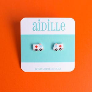May include: A pair of white ambulance stud earrings with red crosses and black wheels. The earrings are displayed on a white and teal card with the word "aidille" in teal and the website address www.aidille.com.