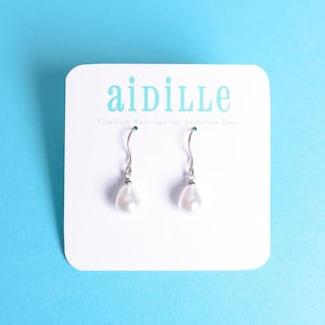 May include: A pair of silver titanium earrings with white pearl drops. The earrings are displayed on a white card with the text "aidille Titanium Earrings for Sensitive Ears".