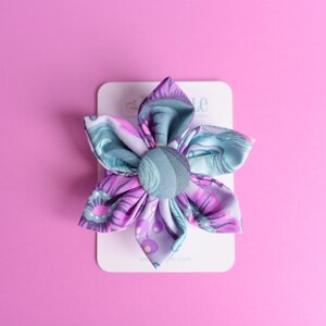 May include: A fabric flower accessory featuring six petals in shades of purple, lavender, and teal with a central fabric-covered button. The floral piece is attached to a white rectangular display card with partially visible text at the top.