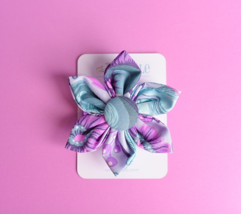 May include: A fabric flower accessory featuring six petals in shades of purple, lavender, and teal with a central fabric-covered button. The floral piece is attached to a white rectangular display card with partially visible text at the top.