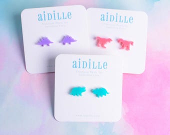 May include: Three cards with the word "aidille" printed on them. Each card features a pair of dinosaur earrings. The earrings are in purple, pink, and turquoise.