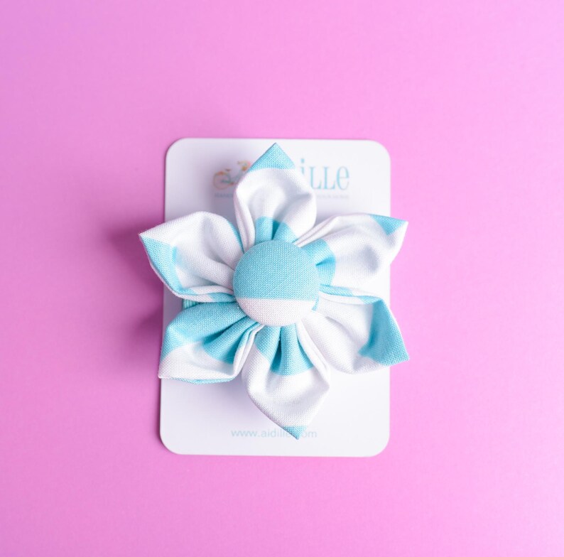 May include: A fabric flower hair accessory featuring six rounded petals with a light blue and white striped pattern. A circular fabric-covered button sits at the center of the flower. The accessory is attached to a white rectangular display card.