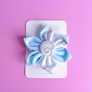 May include: A fabric flower hair accessory featuring light blue petals with a white and grey swirl pattern. The center is a circular fabric-covered button with a matching grey swirl design. The item is attached to a white rectangular display card.