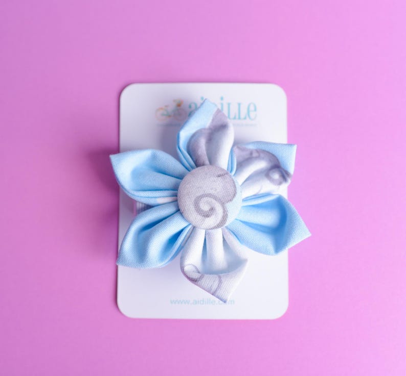 May include: A fabric flower hair accessory featuring light blue petals with a white and grey swirl pattern. The center is a circular fabric-covered button with a matching grey swirl design. The item is attached to a white rectangular display card.
