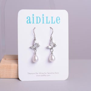 May include: Silver dangle earrings with a pearl drop and a cluster of clear cubic zirconia stones. The earrings are on a white card with the text "aidille" and "Titanium Ear Wires for Sensitive Ears" and "www.aidille.com".
