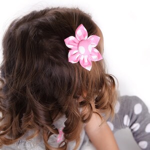 May include: A fabric hair clip featuring a six-petaled flower design in bright pink with white polka dots. The flower has a central fabric-covered button and is attached to a hair accessory base, positioned in dark brown wavy hair.