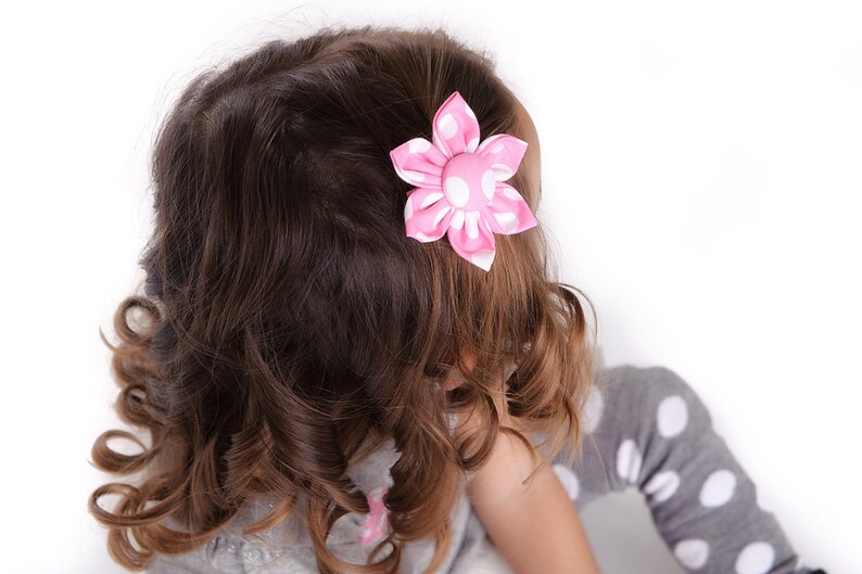 May include: A fabric hair clip featuring a six-petaled flower design in bright pink with white polka dots. The flower has a central fabric-covered button and is attached to a hair accessory base, positioned in dark brown wavy hair.