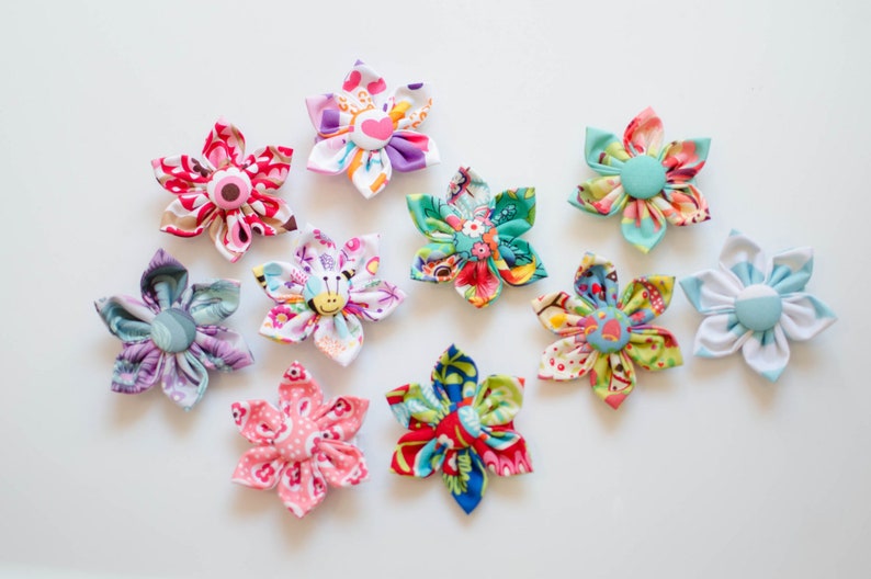 May include: A collection of ten fabric flowers, each featuring six petals and a central fabric-covered button. The flowers display various colorful patterns, including floral, geometric, and animal prints in shades of pink, blue, green, and yellow.
