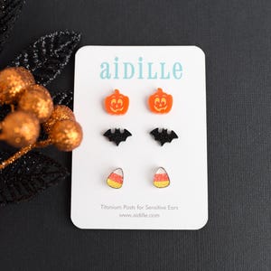 May include: A set of three pairs of Halloween-themed earrings. The earrings are made of acrylic and feature a pumpkin, a bat, and a candy corn design. The earrings are on a white card with the text "aidille" and "Titanium Posts for Sensitive Ears" printed on it.