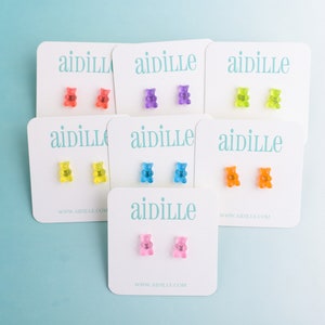 May include: Six pairs of gummy bear stud earrings on white cards with the brand name "aidille" printed on them. The gummy bears are in various colors, including red, orange, yellow, green, blue, and pink.