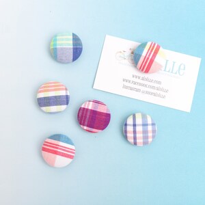 May include: Five circular fabric-covered buttons featuring various plaid patterns in shades of blue, pink, purple, and yellow. A white rectangular business card is present with the text "AIDILLE", "www.aidille.com", "www.facebook.com/aidille", and "instagram @shopaidille".