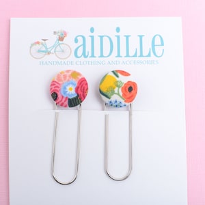 May include: Two floral fabric-covered paper clips. Each clip has a round, colorful floral design. The clips are attached to a white card with the text "aidille HANDMADE CLOTHING AND ACCESSORIES" and a bicycle illustration on a pink background.