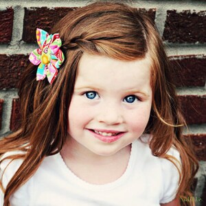 May include: A fabric hair clip featuring a multi-colored floral pattern with pink, yellow, blue, and green hues. The accessory is shaped like a flower with layered petals and is attached to long, wavy auburn hair styled with a side braid.