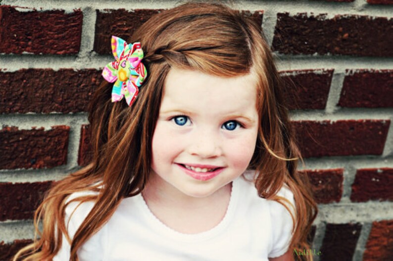 May include: A fabric hair clip featuring a multi-colored floral pattern with pink, yellow, blue, and green hues. The accessory is shaped like a flower with layered petals and is attached to long, wavy auburn hair styled with a side braid.