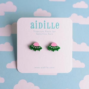 May include: A pair of green glitter UFO earrings with pink domes and white dot accents, displayed on a white card with the brand name "aidille" and the text "Titanium Posts for Sensitive Ears". The background is a light blue pattern with white clouds.