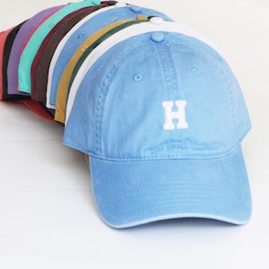 Peut inclure: Une collection de casquettes de baseball de diff&eacute;rentes couleurs, dont rouge, violet, sarcelle, marron, jaune, blanc et bleu clair. La casquette bleu clair &agrave; l'avant porte la lettre "H" brod&eacute;e en blanc. Les casquettes sont dispos&eacute;es en rang&eacute;e, montrant leurs teintes.