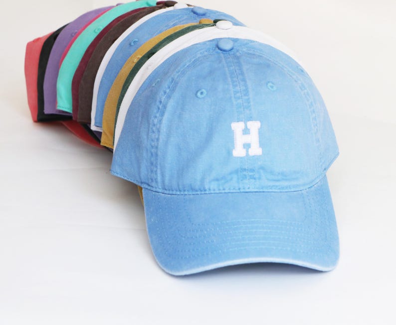 Peut inclure: Une collection de casquettes de baseball de diff&eacute;rentes couleurs, dont rouge, violet, sarcelle, marron, jaune, blanc et bleu clair. La casquette bleu clair &agrave; l'avant porte la lettre "H" brod&eacute;e en blanc. Les casquettes sont dispos&eacute;es en rang&eacute;e, montrant leurs teintes.