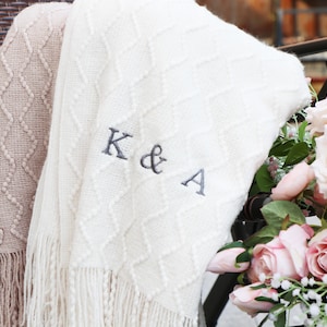 Puede incluir: Manta de punto color crema con un patrón de diamantes y flecos. La manta está bordada con las letras "K & A" en hilo gris. Parcialmente visible una segunda manta de color beige con un patrón similar.