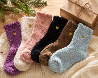 Puede incluir: Un conjunto de seis calcetines mullidos de corte medio en morado, crema, rosa, negro, marrón y azul claro. Cada calcetín presenta un borde con volantes y un pequeño corazón amarillo bordado en el tobillo. Tienen un tejido suave y texturizado.