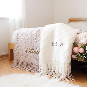 May include: Two decorative blankets on a bed. One is pale pink with the name "Olivia" embroidered in gold. The other is white with "K & A" embroidered in grey. Both blankets feature a diamond pattern and a fringe.