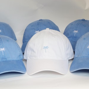 May include: Six baseball caps in a variety of colours. Five are light blue with a white embroidered palm tree logo, and one is white with a light blue palm tree logo. The caps have curved peaks.