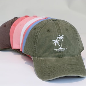 Könnte beinhalten: Eine Auswahl an Baseballkappen in verschiedenen Farben, darunter Olivgrün, Rosa und Braun. Jede Kappe hat einen gebogenen Schirm und ein weißes gesticktes Design mit zwei Palmen und Wellen. Die Kappen sind in einer Reihe angeordnet, um ihre verschiedenen Farbtöne zu zeigen.