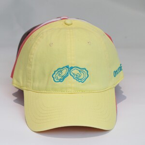 May include: Yellow baseball cap with embroidered blue oyster design. The cap has a curved brim and a button on top. The word "Oysterf" is embroidered on the side. Other caps are visible behind the yellow cap.