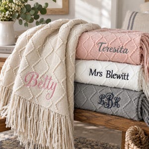 May include: A collection of four soft, textured knit blankets featuring a diamond pattern and fringe edges. The blankets are cream, light pink, white, and grey. They are personalised with embroidered names: Betty, Teresita, Mrs Blewitt, and LBH.