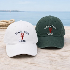 May include: A white baseball cap and a dark green baseball cap, both featuring an embroidered red lobster graphic centered above the text "ACADIA CREW BABE" in navy blue on the white cap and white on the green cap, with a curved brim and button top.