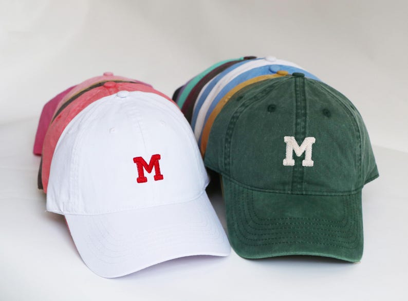 Peut inclure: Plusieurs casquettes de baseball de diff&eacute;rentes couleurs, dont le blanc, le rose et le vert. Chaque casquette pr&eacute;sente la lettre "M" contrastante brod&eacute;e sur le devant. Les casquettes sont dispos&eacute;es sur une surface blanche, mettant en valeur leurs diff&eacute;rentes couleurs et designs.