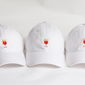 May include: Three white baseball caps with a red cocktail glass embroidered on the front. The cocktail design includes a red drink, a lemon slice, and a straw. The caps are made of a soft fabric and have a curved brim.
