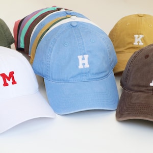 Peut inclure: Plusieurs casquettes de baseball de diff&eacute;rentes couleurs, chacune brod&eacute;e d'une lettre diff&eacute;rente. Les casquettes sont dans des tons de blanc, bleu, marron, jaune et vert, avec des lettres brod&eacute;es blanches ou rouges contrastantes. Les casquettes sont dispos&eacute;es sur une surface blanche.