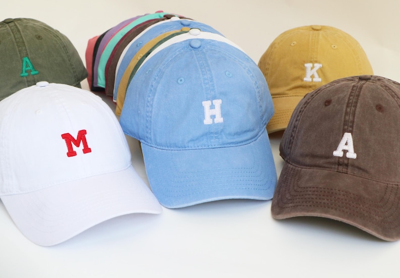 Peut inclure: Plusieurs casquettes de baseball de diff&eacute;rentes couleurs, chacune brod&eacute;e d'une lettre diff&eacute;rente. Les casquettes sont dans des tons de blanc, bleu, marron, jaune et vert, avec des lettres brod&eacute;es blanches ou rouges contrastantes. Les casquettes sont dispos&eacute;es sur une surface blanche.