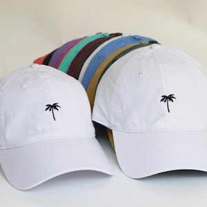 May include: White baseball caps with a black palm tree logo. Several other colored caps are visible in the background, including blue, green, and maroon. The caps are made of a soft material and have a curved brim.