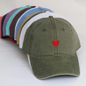 Peut inclure: Une collection de casquettes de baseball de diff&eacute;rentes couleurs, dont vert olive, blanc et bleu. La casquette vert olive au premier plan pr&eacute;sente un petit c&oelig;ur rouge brod&eacute; sur le devant. Les casquettes sont dispos&eacute;es en rang, pr&eacute;sentant leurs diff&eacute;rentes teintes.