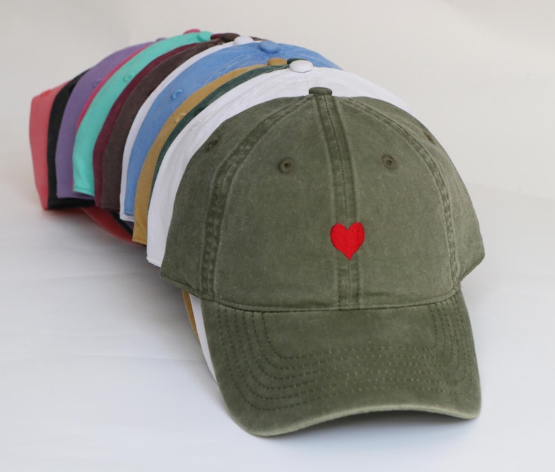 Peut inclure: Une collection de casquettes de baseball de diff&eacute;rentes couleurs, dont vert olive, blanc et bleu. La casquette vert olive au premier plan pr&eacute;sente un petit c&oelig;ur rouge brod&eacute; sur le devant. Les casquettes sont dispos&eacute;es en rang, pr&eacute;sentant leurs diff&eacute;rentes teintes.