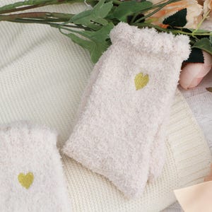 Puede incluir: Un par de calcetines afelpados de color crema con un corazón dorado bordado en el lateral. Los calcetines tienen una parte superior con volantes y se muestran sobre una manta de punto de color crema. El fondo incluye flores y vegetación.