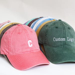 Peut inclure: Plusieurs casquettes de baseball de couleurs vari&eacute;es, dont rose, vert, jaune et bleu. La casquette rose au premier plan porte un "C" blanc brod&eacute;. La casquette verte affiche "Custom Logo" en blanc.