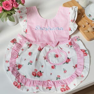 May include: A pink and white floral apron with a matching pot holder. The apron features a pink gingham top and a white skirt with red rose patterns. The name "Shoshana" is embroidered in blue on the top.
