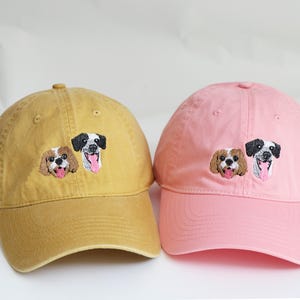 May include: Two baseball caps, one mustard yellow and one pink, each embroidered with two dog faces. The dogs have brown, black, and white fur, with pink tongues. The caps have a curved peak and adjustable strap.