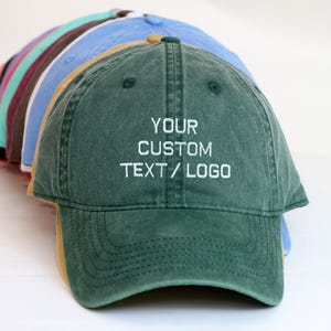 Può includere: Una fila di cappellini da baseball in vari colori, tra cui verde, blu e rosa. Il cappellino anteriore è verde scuro con la scritta "YOUR CUSTOM TEXT / LOGO" ricamata in bianco. I cappellini sono disposti in un leggero arco.